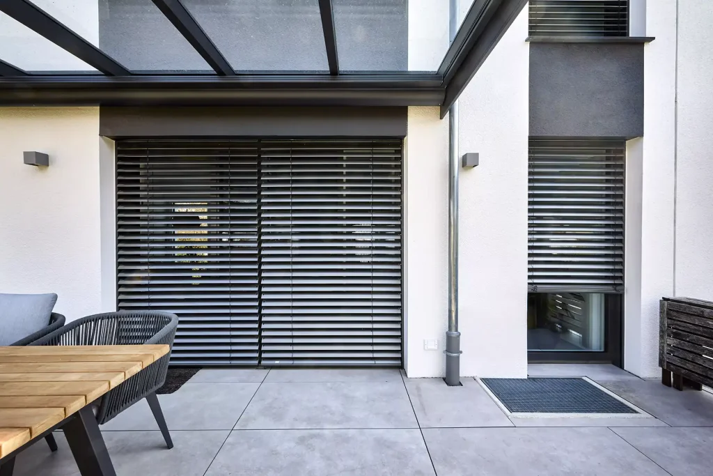 Moderne woning met zwarte buitenjaloezieën onder een glazen terrasoverkapping.