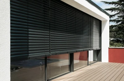 Donkergrijze ROMA buitenjaloezieën voor brede ramen van een moderne woning met wit stucwerk en houten terras.