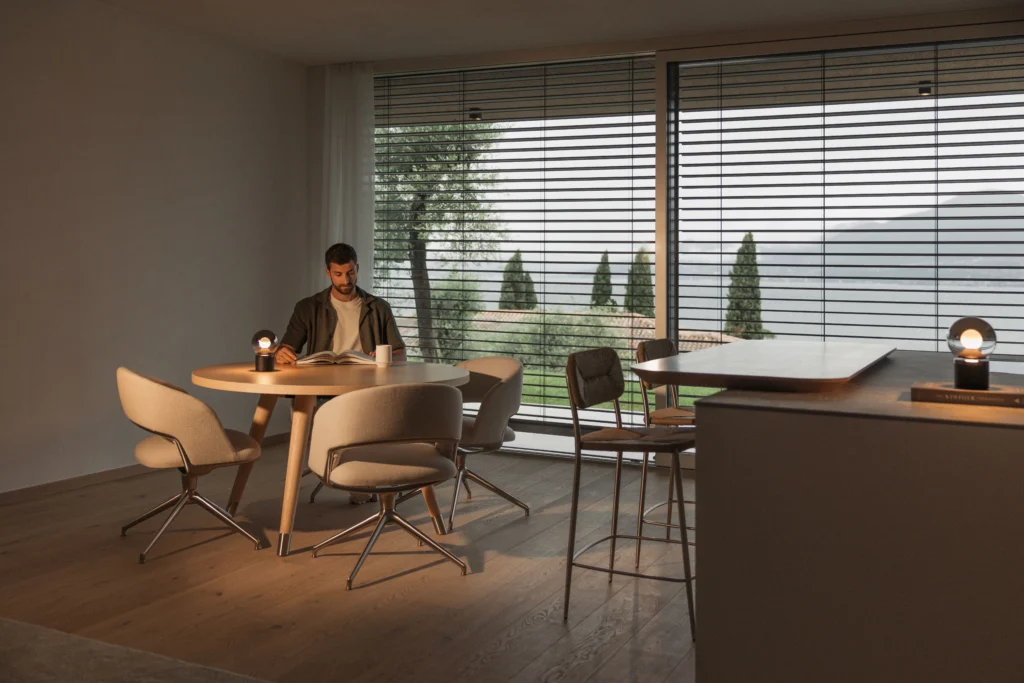 Man leest aan een eettafel in een moderne woonkamer met buitenjaloezieën voor grote ramen. | Zonwering.nl