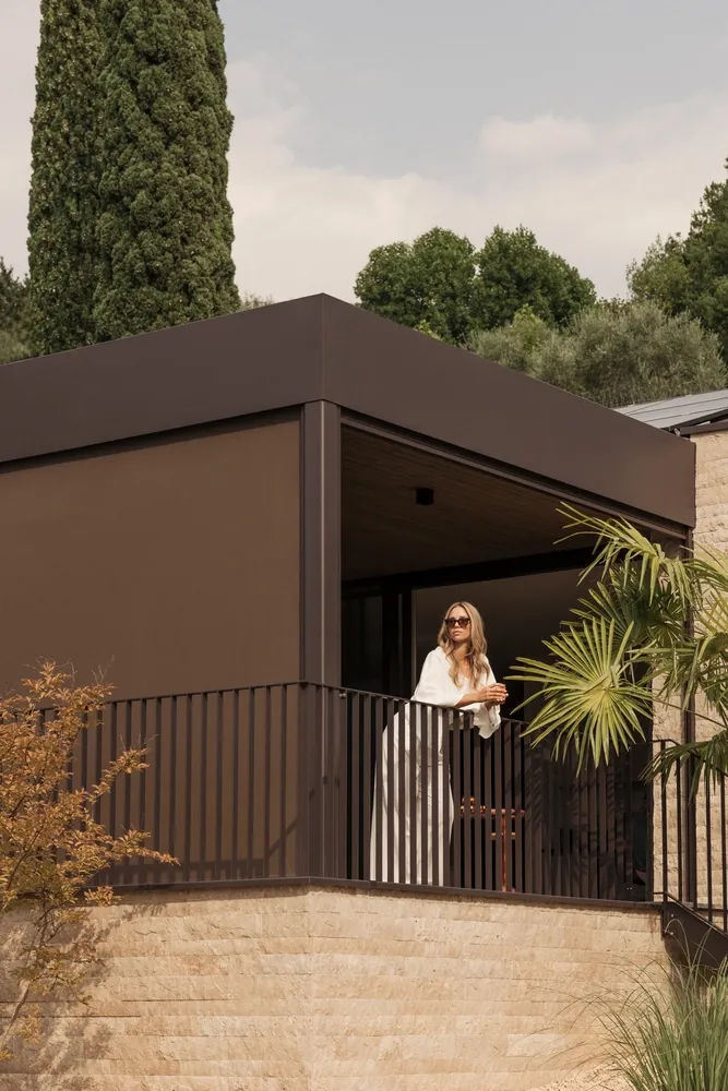 Vrouw staat op een modern overdekt balkon met bruine screens en uitzicht op een groene tuin. | Zonwering.nl