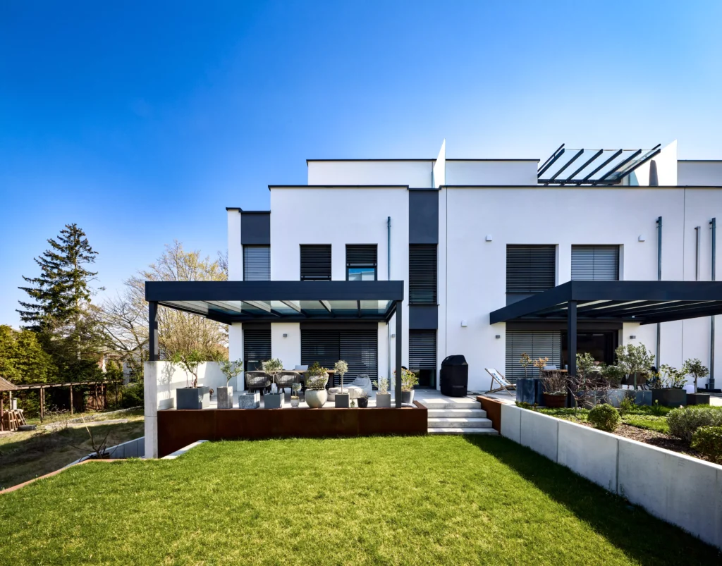 Moderne witte woning met zwarte buitenrolluiken, terrasoverkappingen en een groene tuin. | Zonwering.nl