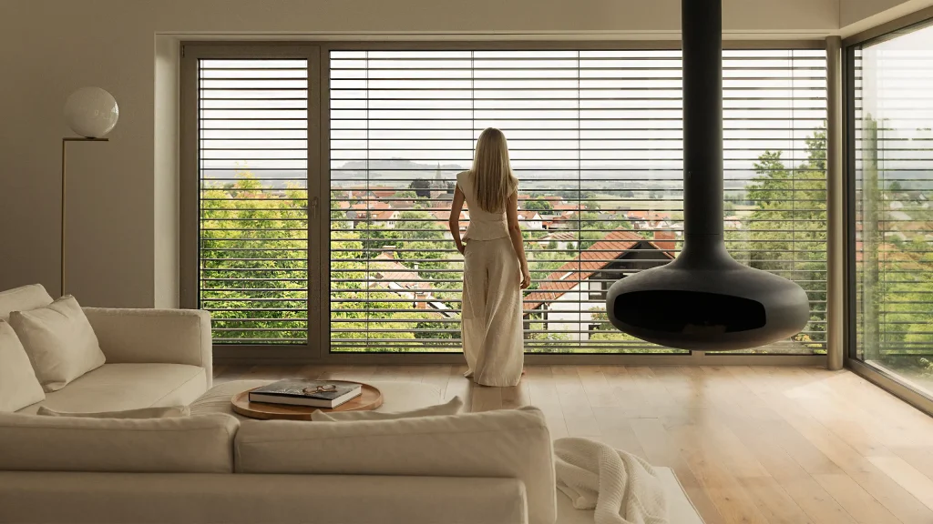 Vrouw kijkt vanuit een moderne woonkamer door grote ramen met buitenjaloezieën naar buiten. | Zonwering.nl
